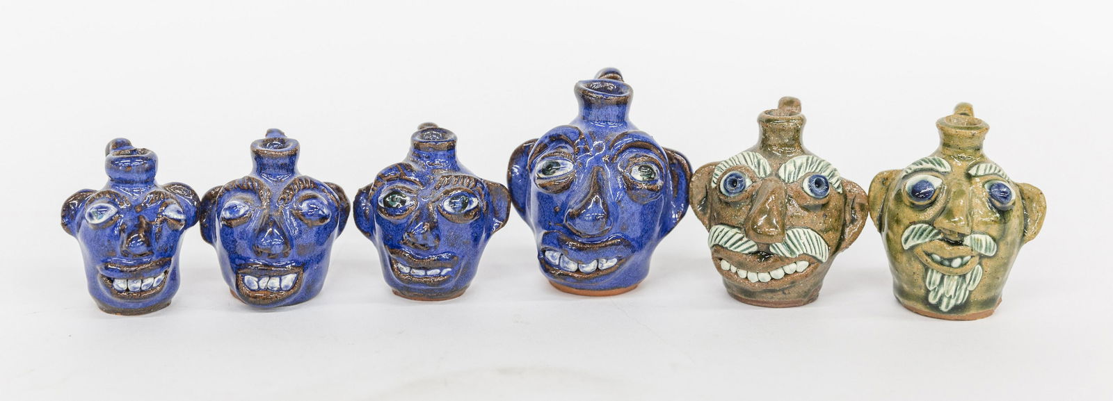 Six Albert Hodge Miniature Face Jugs: Including four in blue alkaline glaze on redware, from 2 1/4 to 3" high; and two brown alkaline glazed stoneware with applied porcelain eyes and teeth, stamped on backs and bases, Albert Hodge Vale N.
