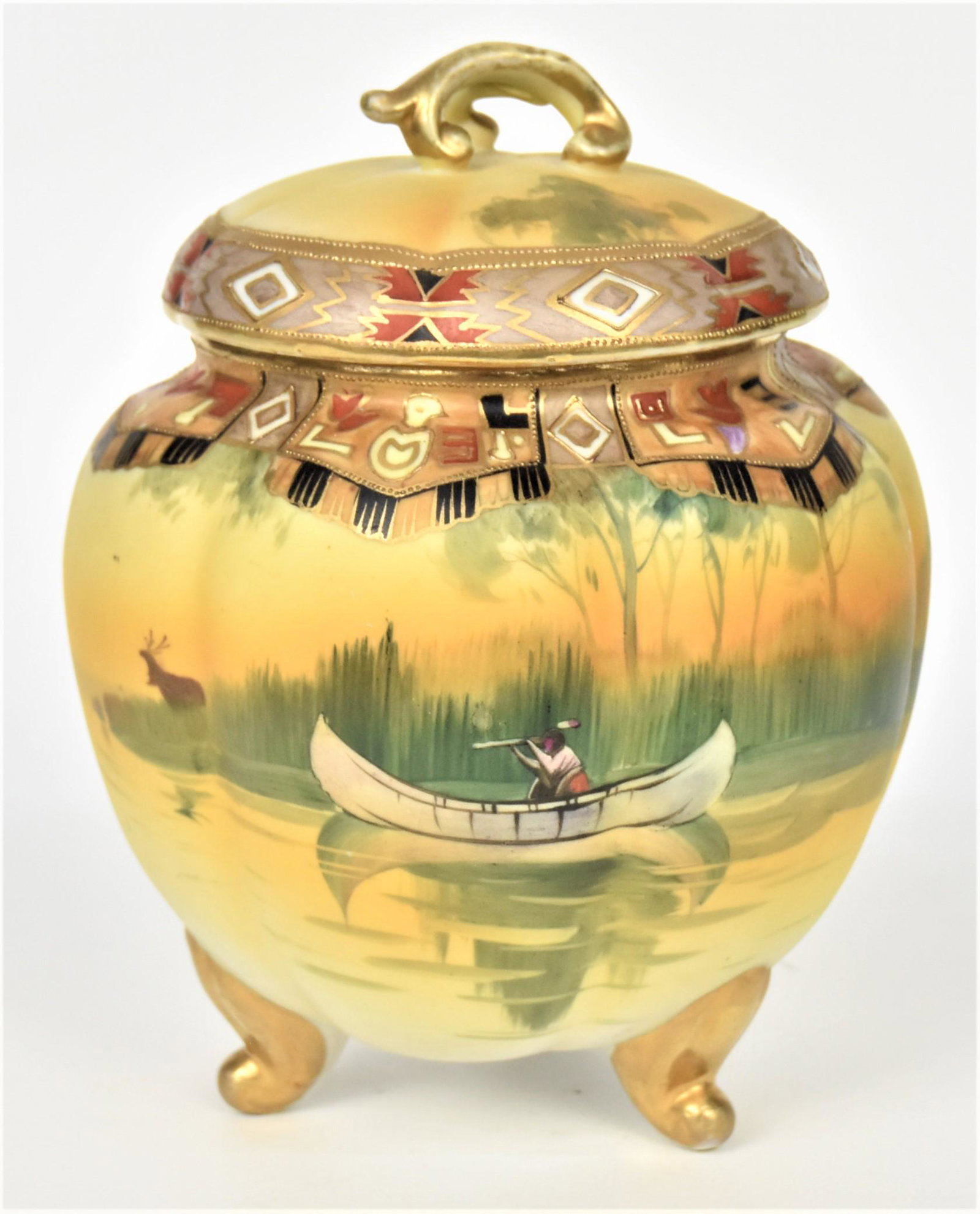 Nippon Scenic Cracker Jar With American Indian: American Indian in a canoe, hunting a stag at dawn. Triple scrolled feet loft a melon ribbed container. Painted Indian designs on shoulder and on cover. M in wreath backstamp 8 3/4" tall, 7" diameter.