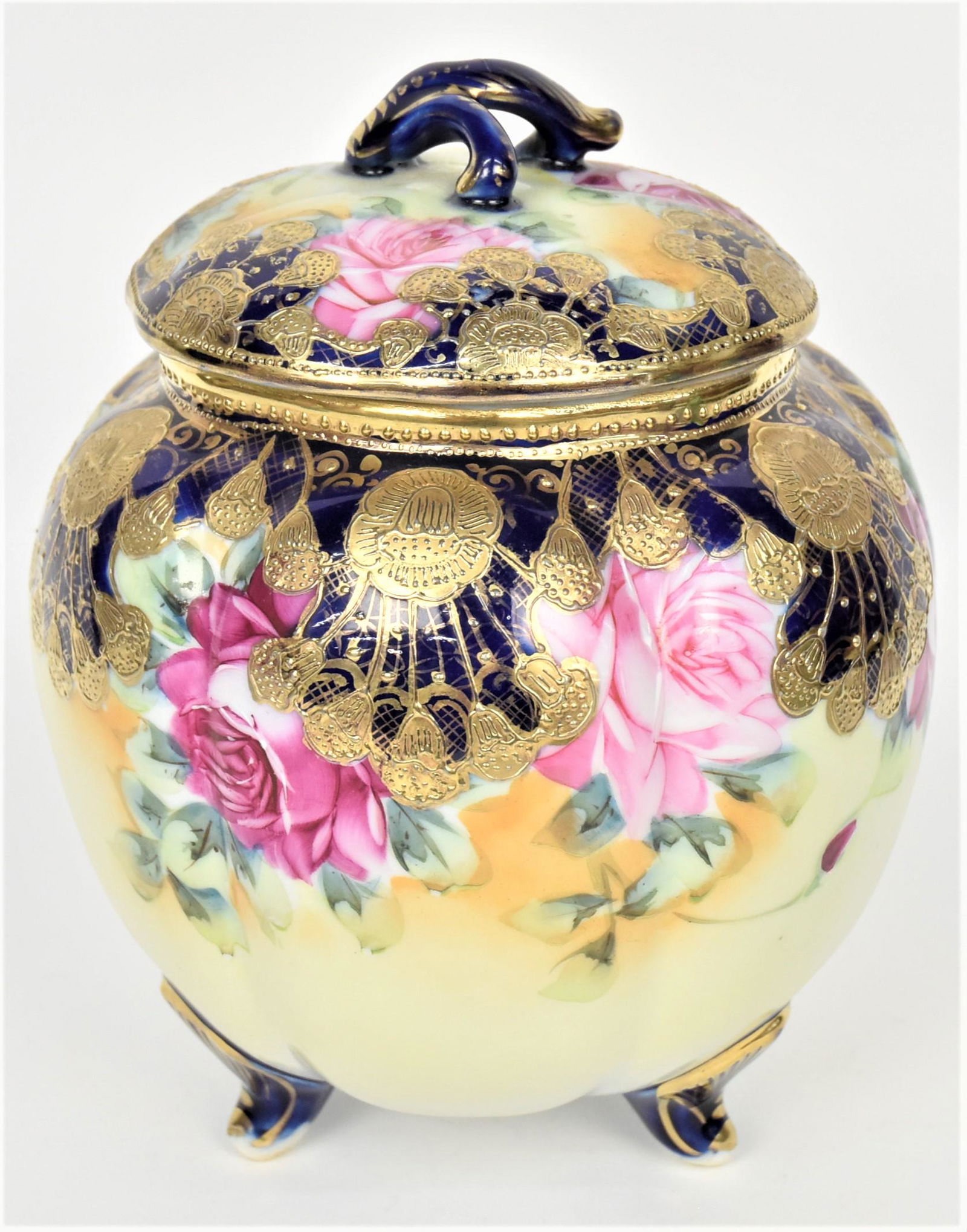 Nippon Cobalt and Gold Cracker Barrel: A melon rubbied body surrounded by ruby and pink roses. Enhanced with raised gold floral, tri-footed. Maple leaf logo, 8" tall. Cover broke and repaired.
