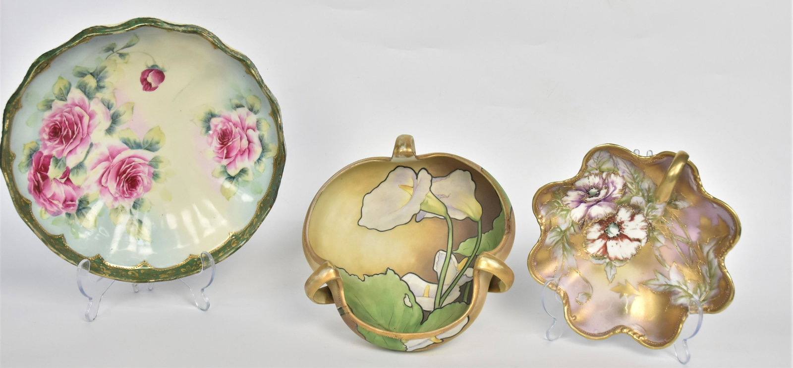 Three Nippon Hand Painted Pieces: A tri-form handled bowl, 3" x 8"D with large lilies & foliage, a handled nappie, 2 1/2" x 7/12dia with poppies and gold highlighting, a center bowl with pink rose boquet, 3" x 10" dia. Small repair to
