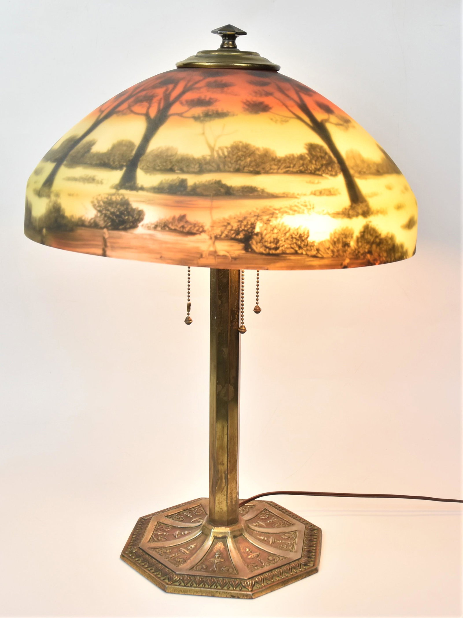 Reverse Painted Table Lamp: Domed 16" diameter glass shade with forested landscape painting, mounted on brass plated cast iron base with neo-classical esign, 23" high. Condition wear to plate and crack in shade.