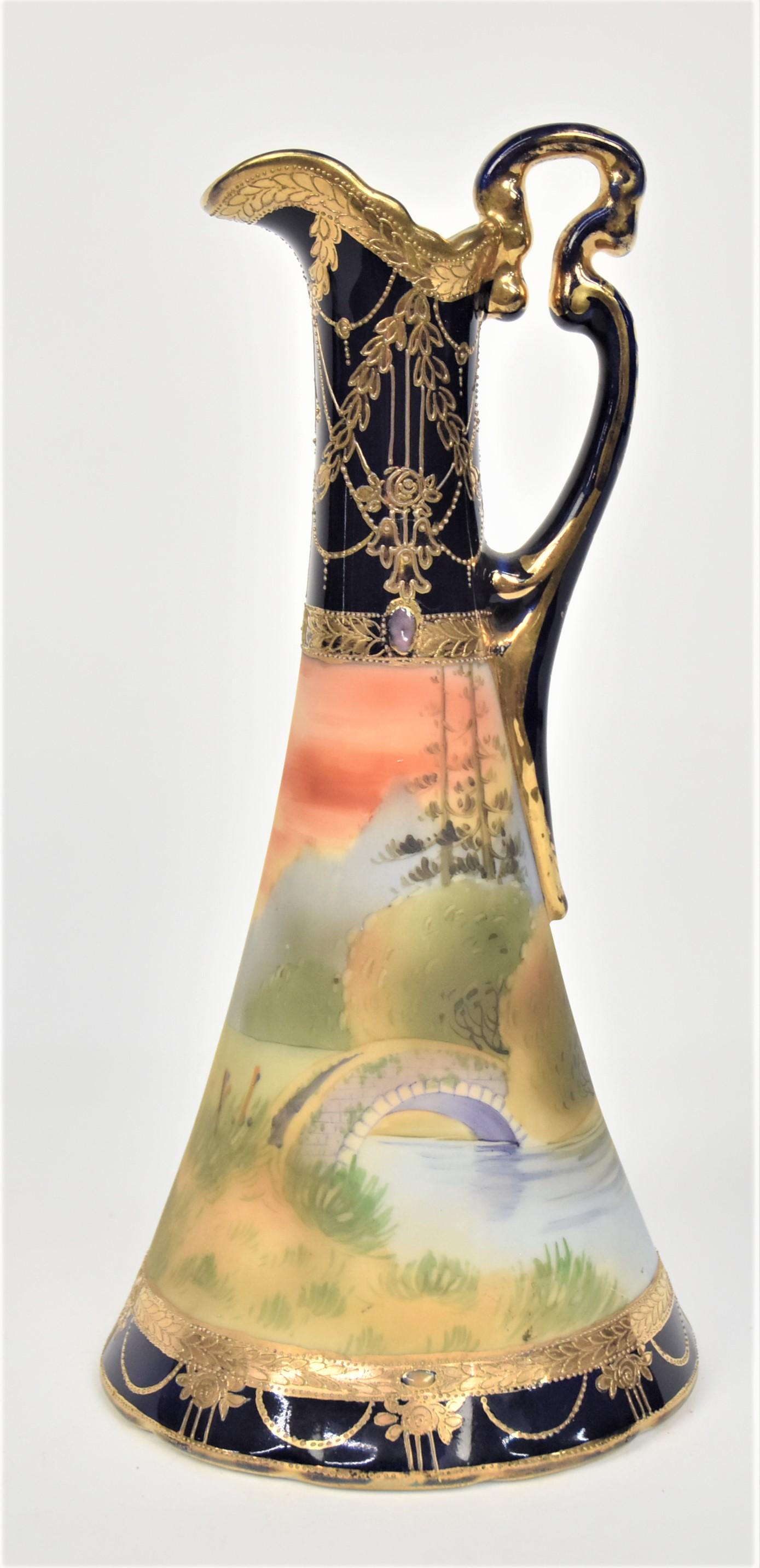 Nippon Cobalt Blue & Jeweled Landscape Ewer: A rock bridge spans an inlet lake at eventide. Raised gold enhancements. Scroll handle. 10 1/4”T. 5 1/4”W base. Very good condition. Green mark.