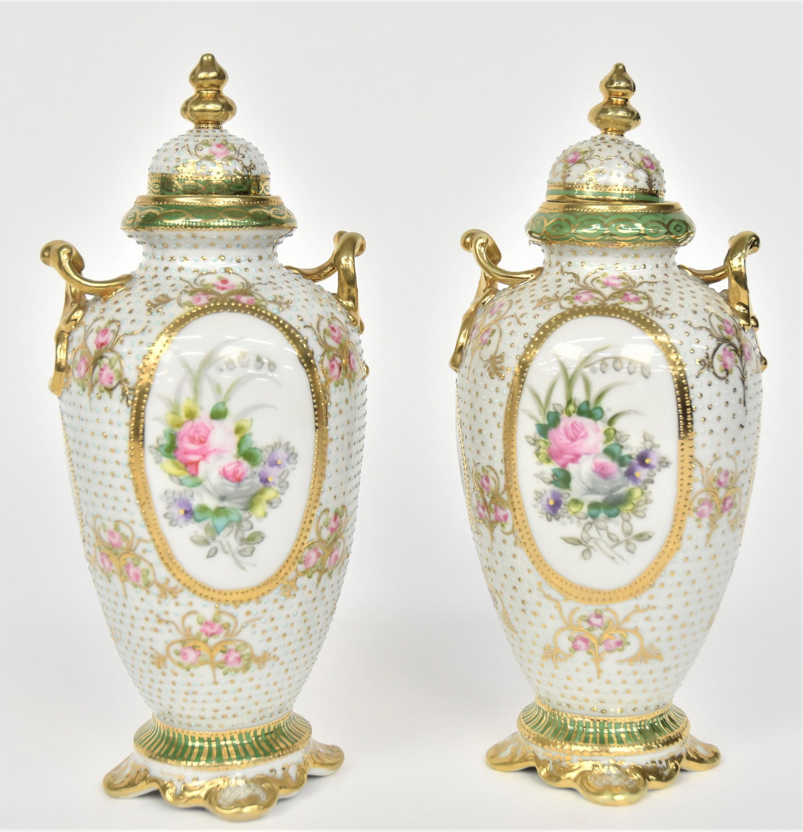 Pair of Nippon Gold & Enamel Beaded Urns With Covers: Oval windows reveal bouquets of colorful garden flowers. Baby roses scatter the body. Fancy scroll handles. 9 3/4”T. 4”W body. Attractive pair. Very good condition. Blue marks to each.