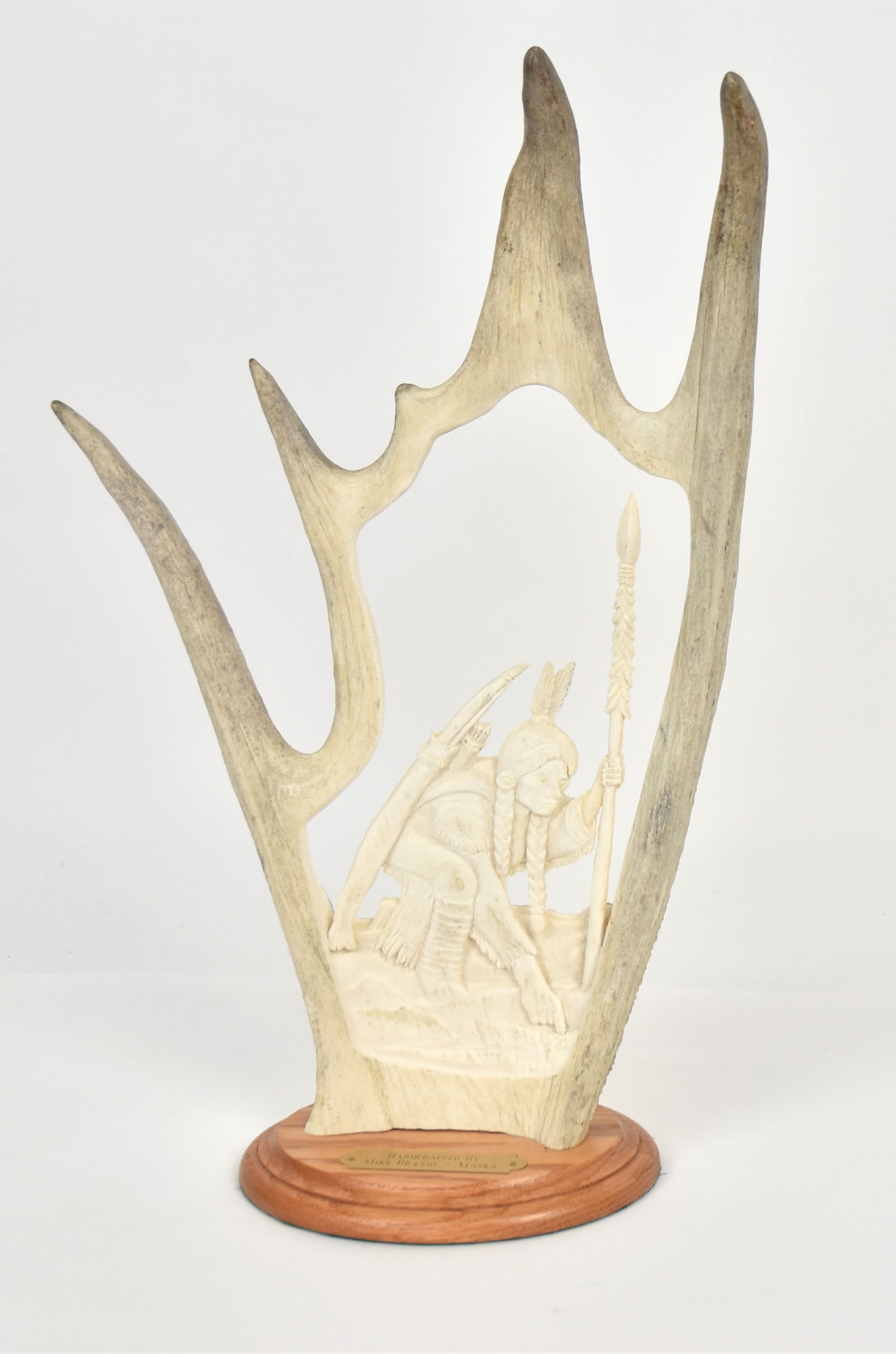 Mike Brandt Moose Antler Sculpture: Cut-out and carved antler with Northern Plains Native American hunter crouching down and pointing to deer hoof print, mounted on oval oak base with brass escutcheon reading: Handcrafted By/Mike Brandt