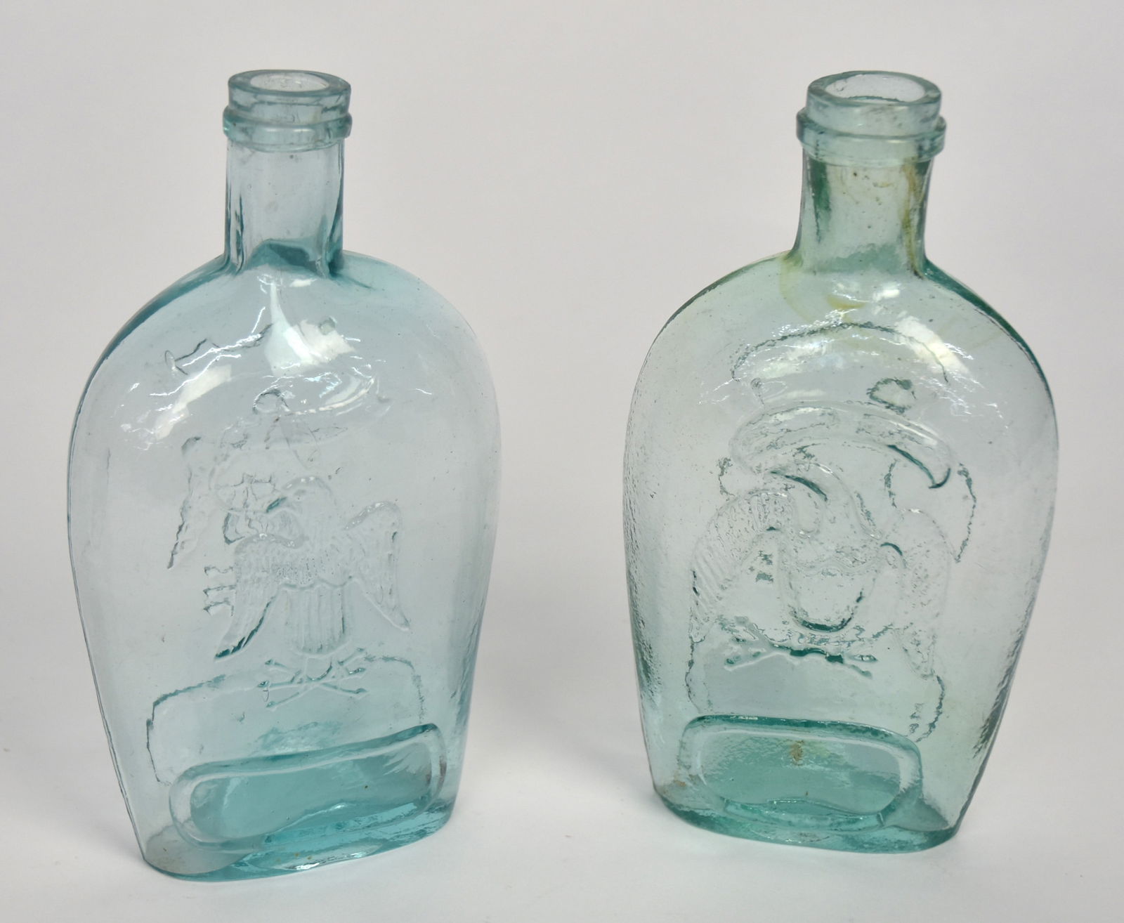 Two Aqua Eagle Flasks: Both different by same motif with spread wing U.S. shield breasted eagle, ribbon banner in beak, clutching laurel branch and arrows in talons with oval escutcheon below on each side, applied ring just