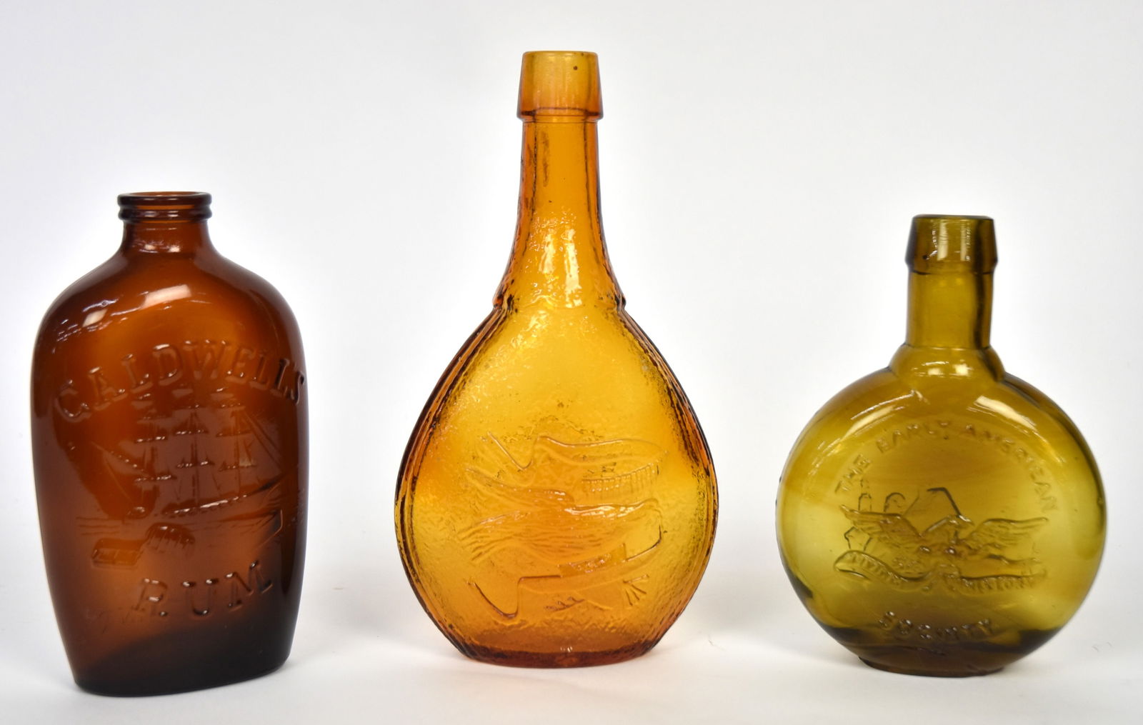 Three Later Colored Glass Flasks: Including deep amber Caldwells Rim with three masted ship unloading barrels of rum, reverse with One Pint and Federal Law text, 7 1/4" high; plus olive green The Early American Society Eagle flask wit
