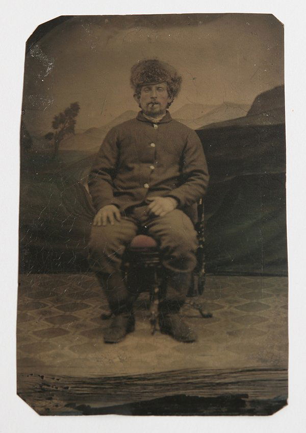 UNUSUAL INDIAN WAR TINTYPE PHOTO: Of a seated soldier in uniform wearing cavalry type boots to the knees, 4 button frock coat and winter fur cap. Seated in Victorian chair with cigar in mouth, in front of a painted landscape backgroun