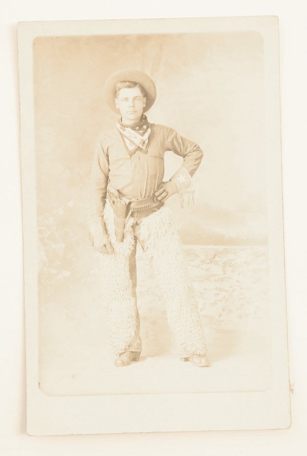 REAL PHOTO POSTCARD OF ARMED COWBOY: 3.5” x 5.5” wearing broad brimmed hat, gauntlets with embroidered stats, sheep skin chaps and holster with Colt type revolver.