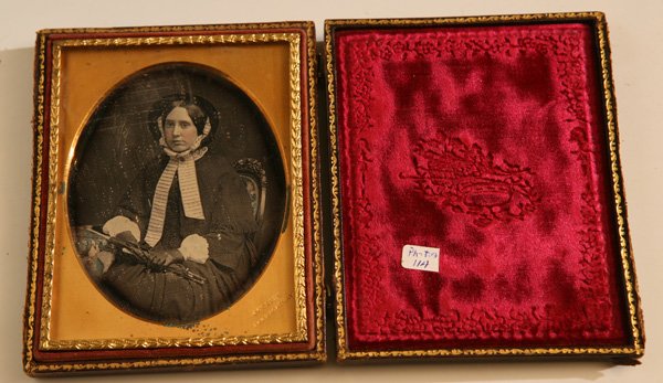 QUARTER PLATE DAGUERREOTYPE BY J. GURNEY: Of a lovely young lady in winter bonnet and outfit and holding an umbrella with folding handle in her gloved hands, beautifully hand tinted. Marked on brass mat “J. Gurney/349 Broadway” and housed