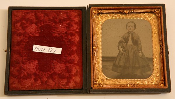 WONDERFUL CIVIL WAR AMBROTYPE OF GIRL & DOLL: A sixth plate in full patriotic case, full length portrait of a little girl in dress with darker piping, typical of this war era, sort of copying soldiers uniforms. She holds her fully dressed black h