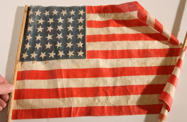38 STAR U.S. CENTENNIAL PARADE FLAG: Printed linen and glazed with canton patter of 7-6-6-6-6-7, mounted on original turned wood stick, 12.25 x 17.75” with 24” stick.