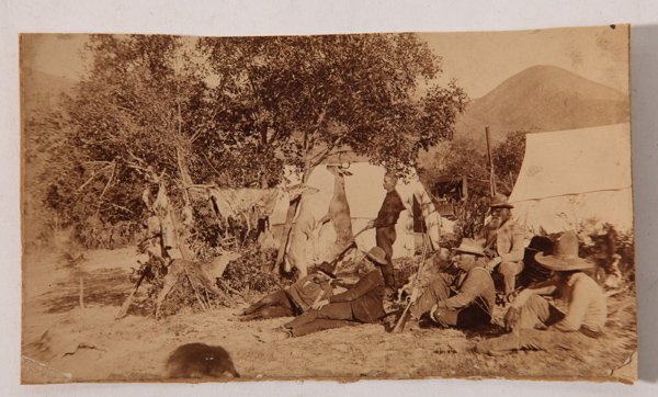 Albumen Photo of Western Hunting Party: A 3.4 x 5.8 inch albumen photograph of a western hunting parties camp with four Anglo men, one African American and one Mexican man, most seated on ground, but one standing with rifle in hand, one sea