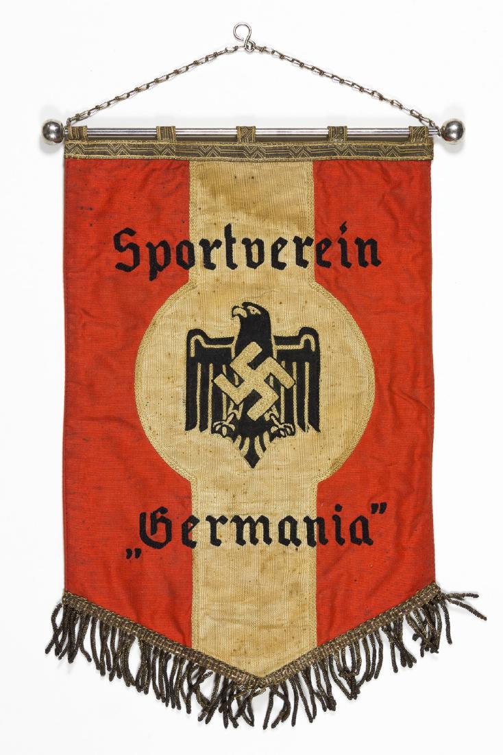 SPORTVEREIN 'GERMANIA' TABLE BANNER: Small Third Reich table banner, 7" x 10", constructed of red and taupe rayon and showing an embroidered eagle with folded wings and a mobile swastika at its breast in black fabric and taupe thread, su