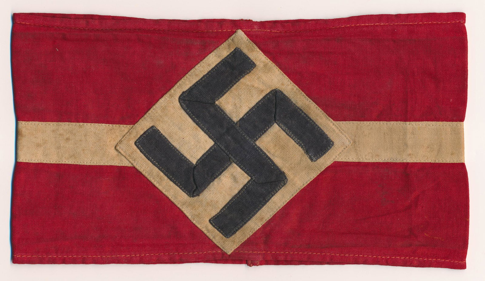HITLER YOUTH ARMBAND: HITLER YOUTH ARMBAND Red cotton armband, approx. 7' x 3 3/4', with an applied white band and an applied white diamond and mobile swastika. Bears a very faint HJ rubber stamp on the face. Somewhat soil