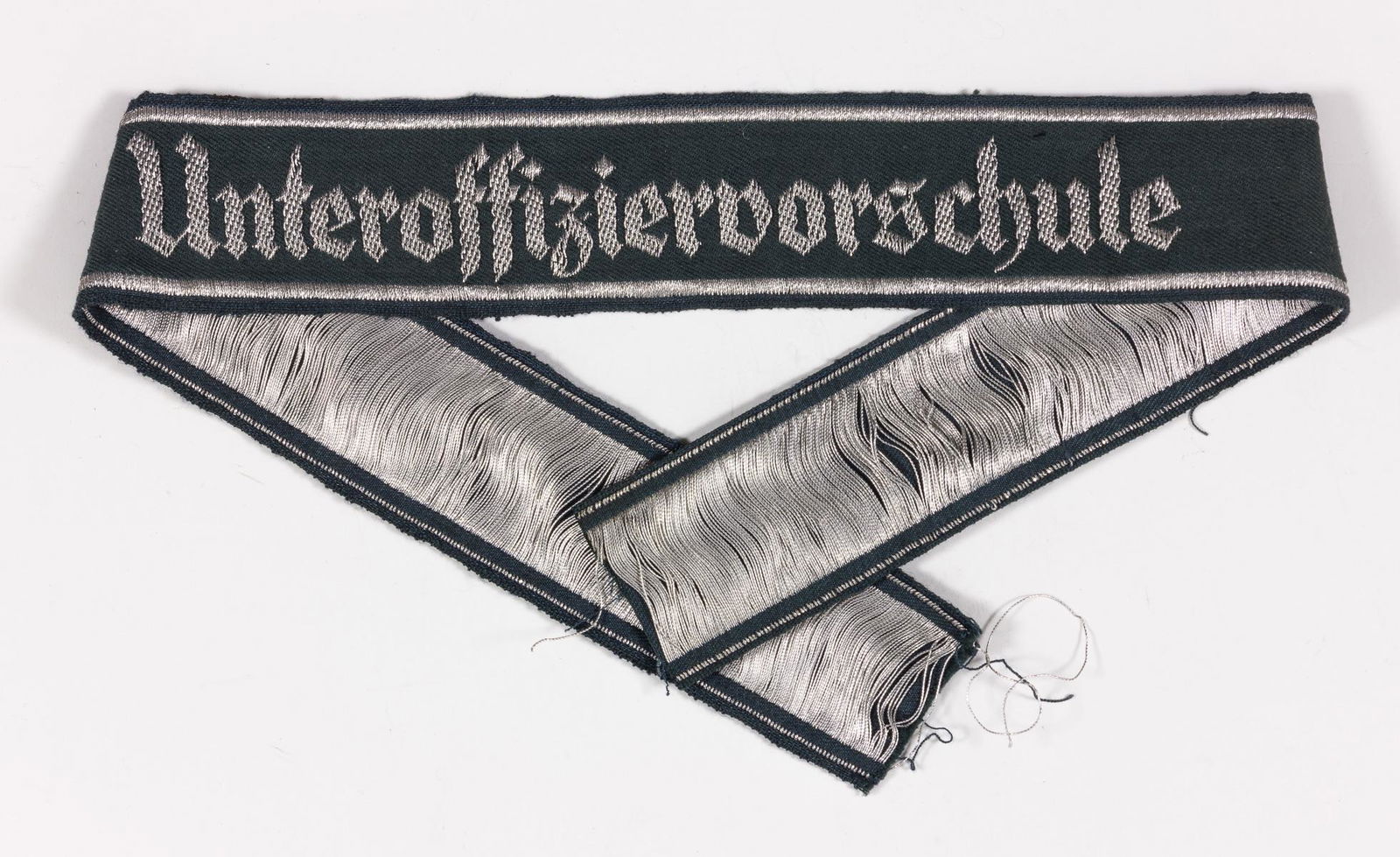 NON-COMMISSIONED OFFICER'S SCHOOL CUFF TITLE: BeVo-style embroidered cuff title, 17.5 in. x 1.25 in., bearing the legend 'Unteroffiziervorschule' in silver thread on a dark green background bordered with bands of silver thread, indicating wear by