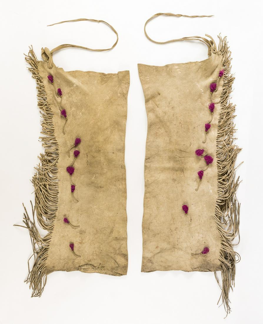INDIAN FRINGED DEERSKIN CHAPS: Pair of antique Indian deerskin chaps, 30' long, showing obvious use. The chaps each bear eight decorative purple wool tufts with brass beads hanging in a vertical row, beside the fringe which runs th