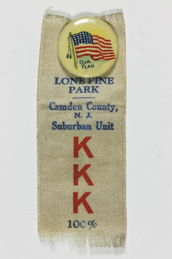NEW JERSEY KU KLUX KLAN BADGE: Celluloid-covered pinback badge, 3/4' dia., depicting an American flag and marked on the verso as being manufactured by James Spencer & Co. of Philadelphia. Attached to the badge is a 3' x 1' white ri