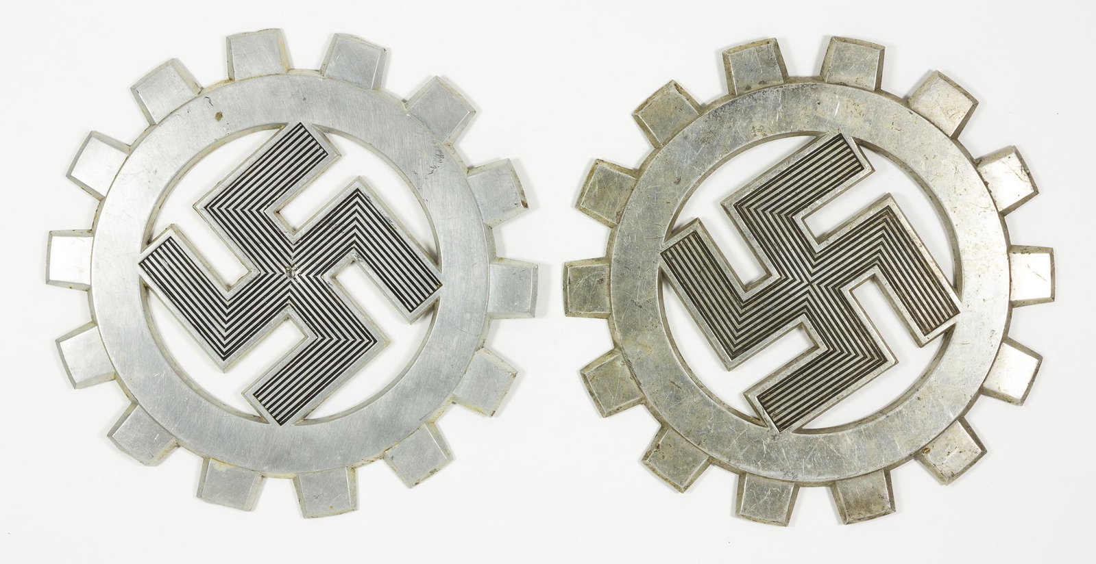 D.A.F. PLAQUES (2): Lot of two aluminum plaques, both approx. 8 in. dia., showing the emblem of the"Deutches Arbeitfront"Â, a mobile swastika within a gear. Both bear minor wear and bumps to some of the gear's prongs, o