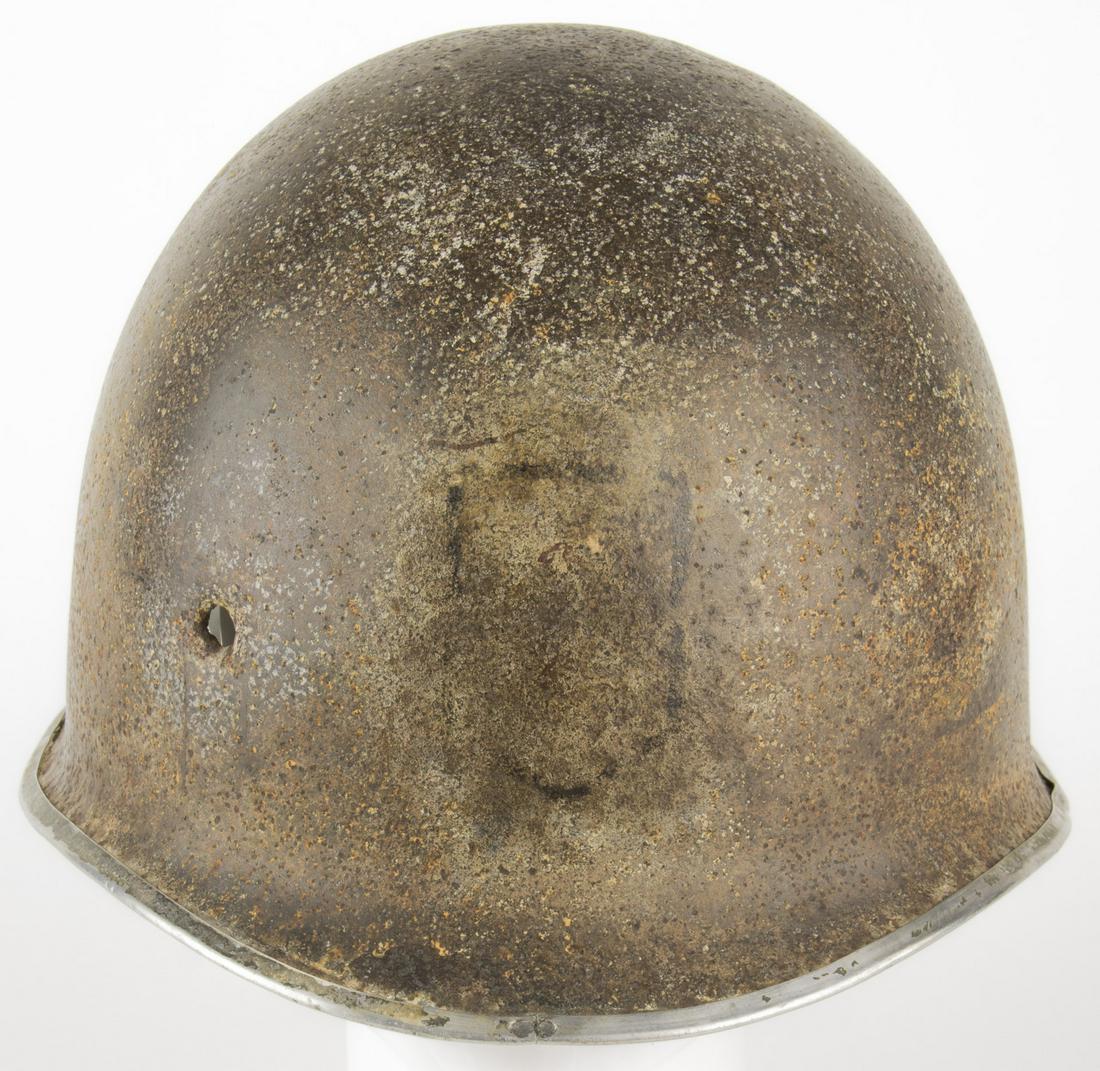 BATTLE-DAMAGED M1 HELMET WITH DIVISIONAL INSIGNIA: An evocative relic purportedly recovered near Omaha Beach, a United States Army M1 steel helmet shell, with fixed strap bales, a seam at the front, and the paint of the rim flaking away to reveal the 