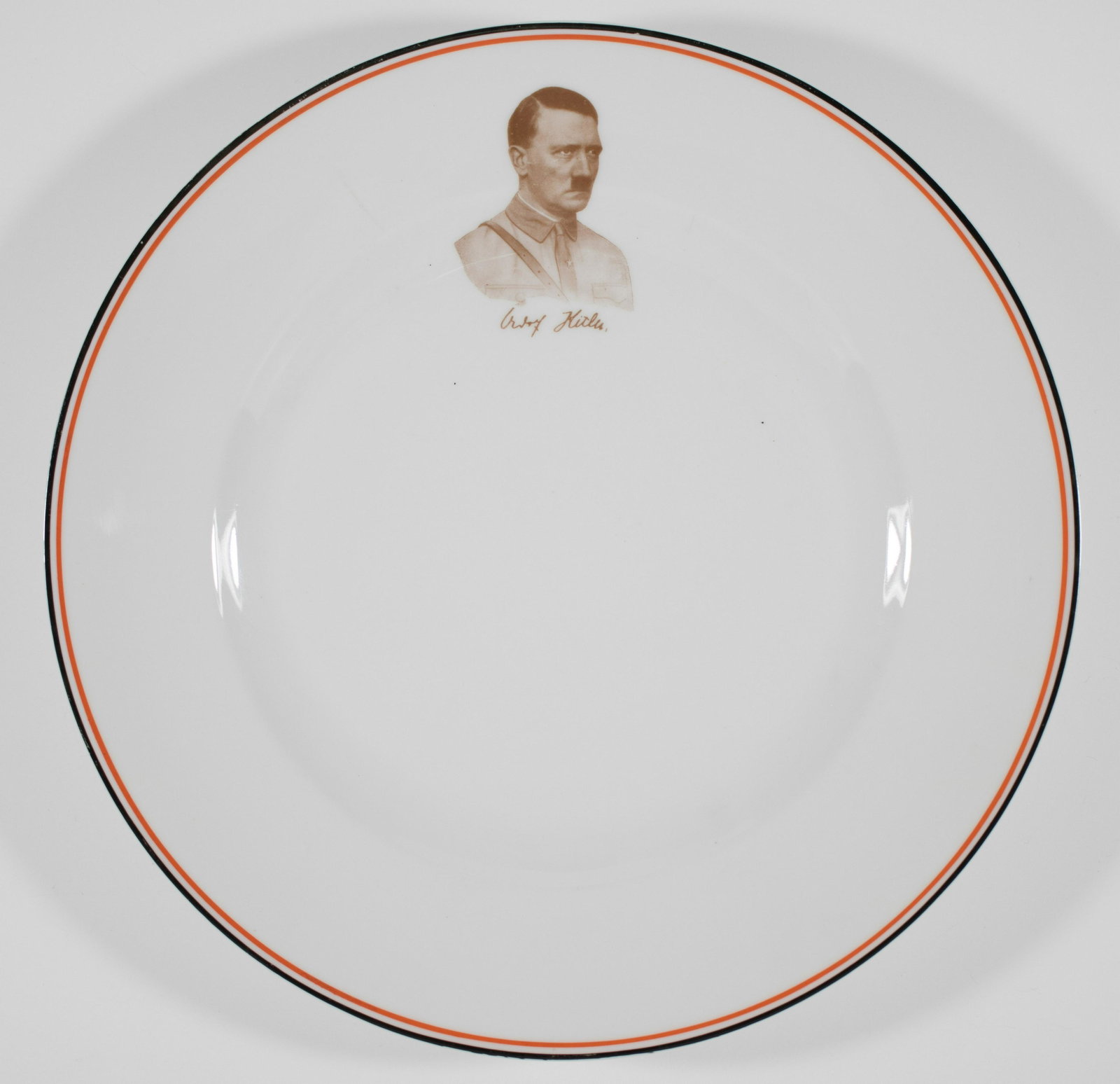 ADOLF HITLER COMMEMORATIVE PLATE: German porcelain plate 9.5 in. diameter, bears a sepia chest, up image of Adolf Hitler in Brownshirt uniform with his facsimile signature beneath. The plate is rimmed in red, white and black, the Germ