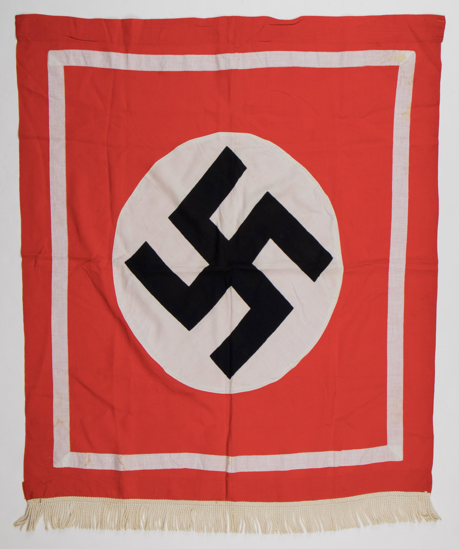 NAZI PARTY PODIUM BANNER: Vibrant Nazi Party podium banner, 28 x 32 in., constructed of red cotton fabric with a sewn-on white disk bearing a printed mobile swastika at its center, also bearing a white sewn-in border. Two whit