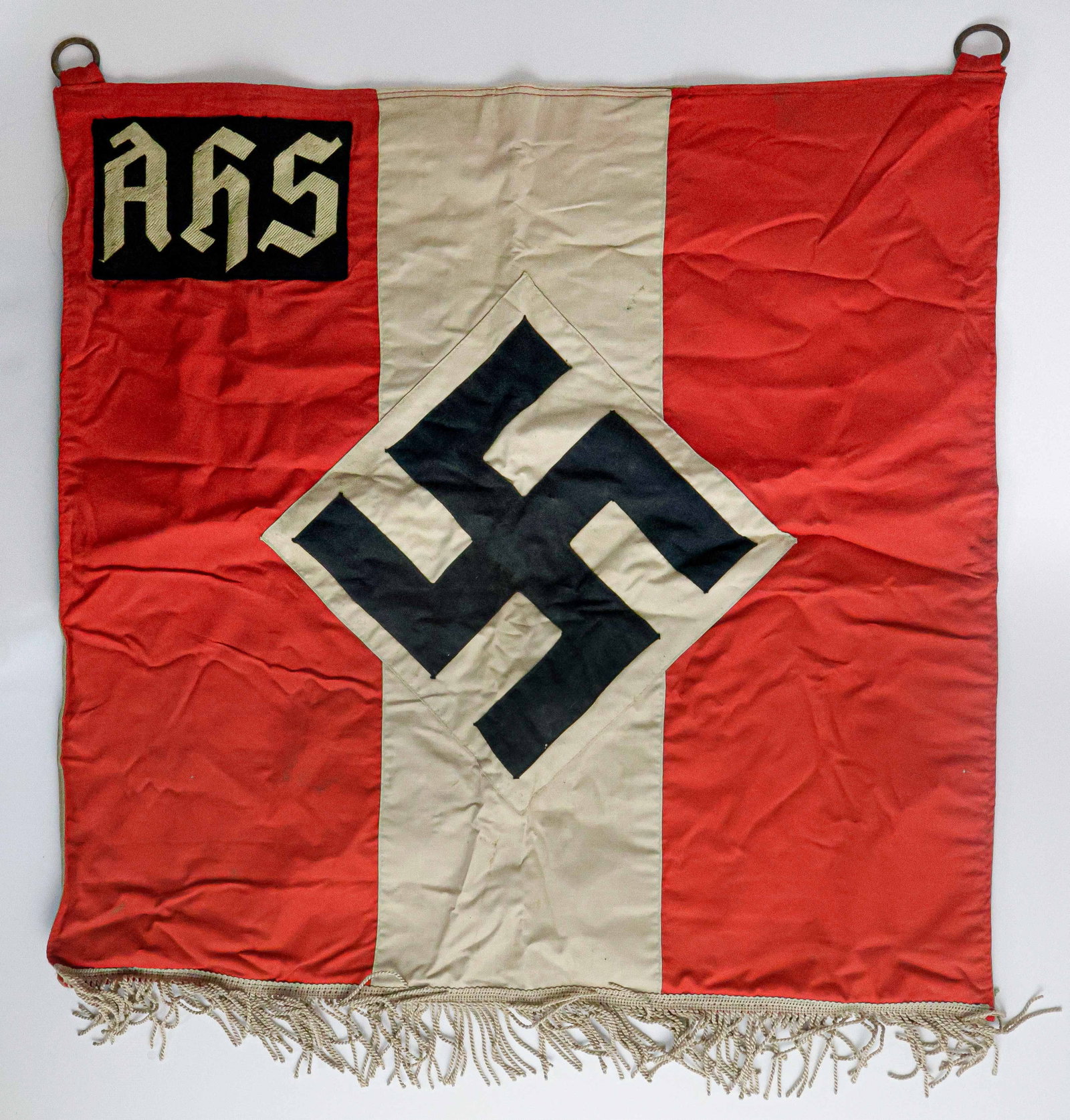 ADOLF HITLER SCHOOL BANNER: A cotton, one-sided, multipiece construction NSDAP banner, 24 x 24 in., with black mobile swastika at center, ‘AHS’ [Adolf Hitler-Schule] at top left, and satin cord fringe at bottom. With