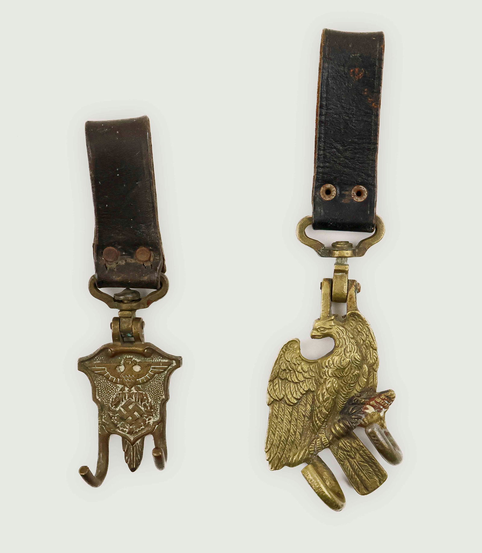 HITLER YOUTH DRUM HANGERS (2): Set of two Hitler Youth drum hangers with leather belt loops and of cast brass construction. The first, 6.5 in. long, has an ornate shield base with scalloped sides and a raised edge border, encompass
