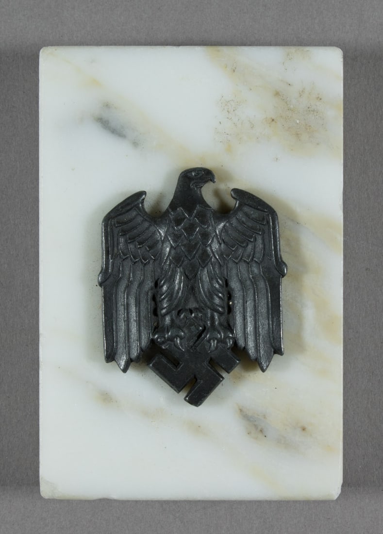 EAGLE AND SWASTIKA PAPERWEIGHT: Desk paperweight, 2 x 3 x .75 in., white marble with a dark metal national eagle with swastika affixed to the upper surface, the green felt on the bottom, obviously replaced. Fine.