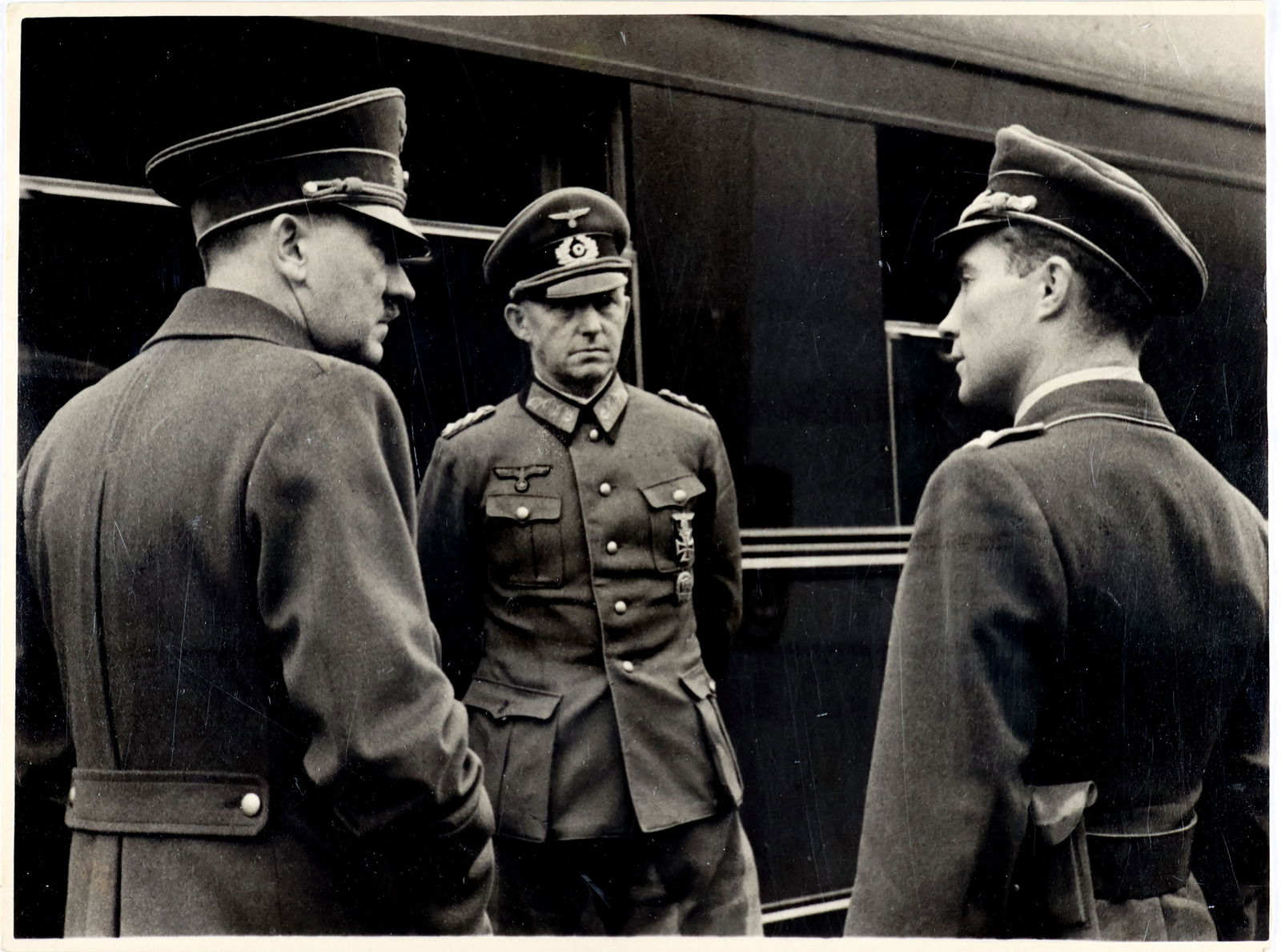 HEINRICH HOFFMAN HITLER PHOTOGRAPH: Original Adolf Hitler photograph, 9 x 7 in. b/w, showing a profile view of Hitler as he speaks with two Nazi officials, all standing before a train. Purple press stamp on the verso reads: ‘Press