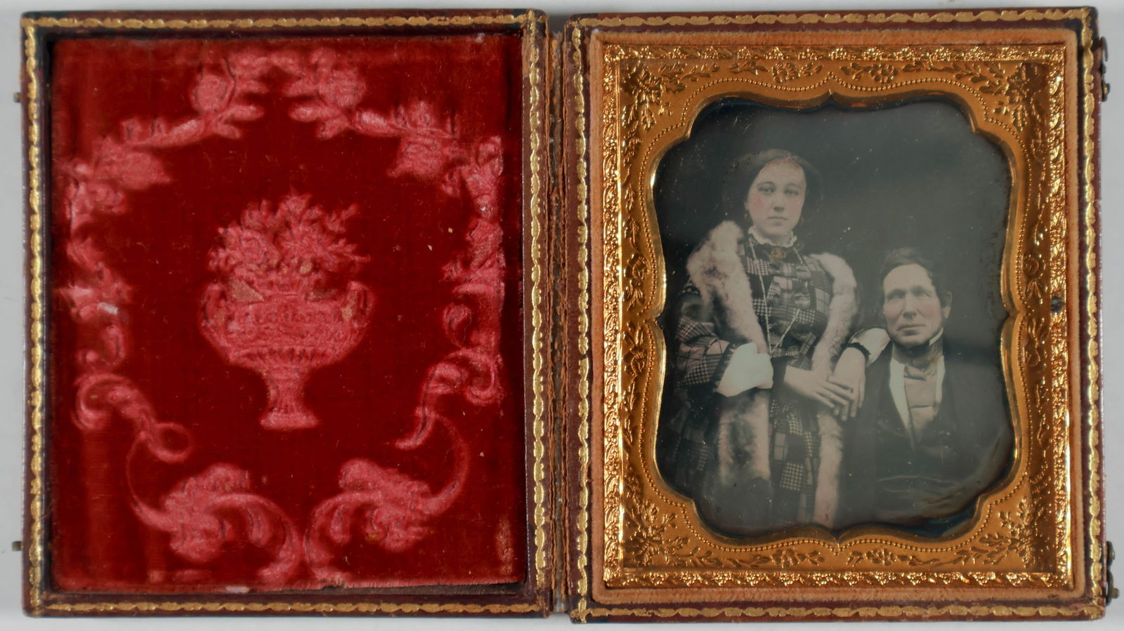 DAGUERREOTYPE OF A WEALTHY COUPLE: A daguerreotype, 2 x 2.5 in. (sight), of a man and his wife who is wearing an elaborate coat with a fox fur and a hand tinted gold necklace. Within an intact case with broken hinge, otherwise fine.