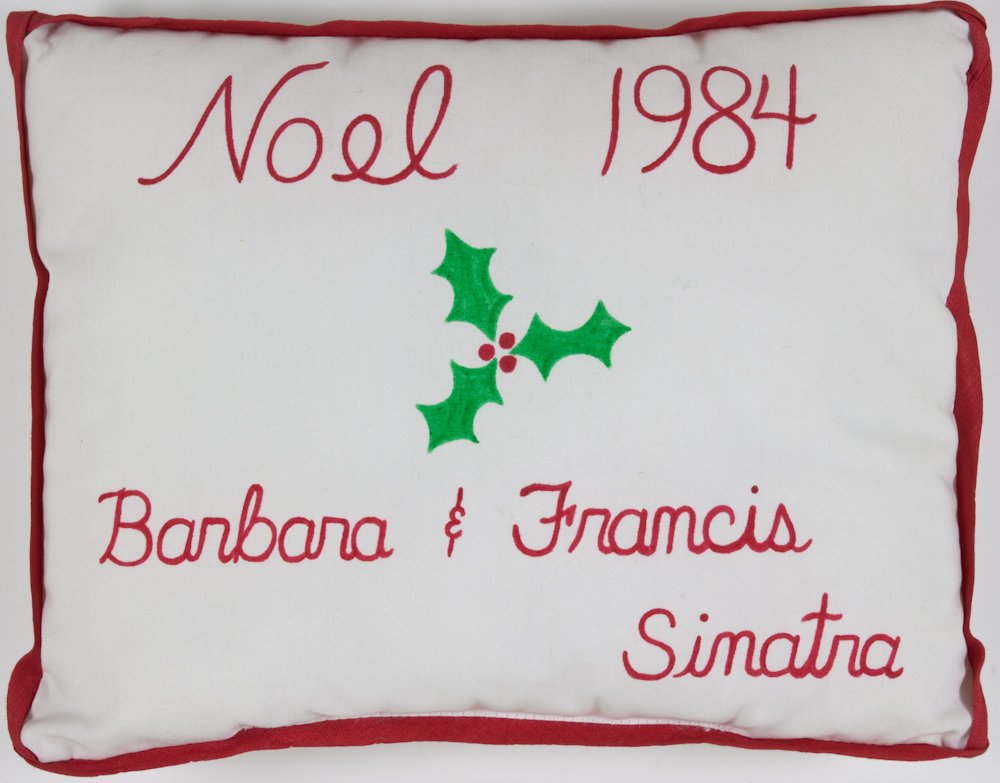 FRANK SINATRA'S CHRISTMAS GIFT: A great Sinatra relic, a 9 1/2" x 7" white pillow with red trim, bearing red hand-printed lettering: "Noel 1984 Barbara & Francis Sinatra". At center appears a small sprig of holly with three red berr