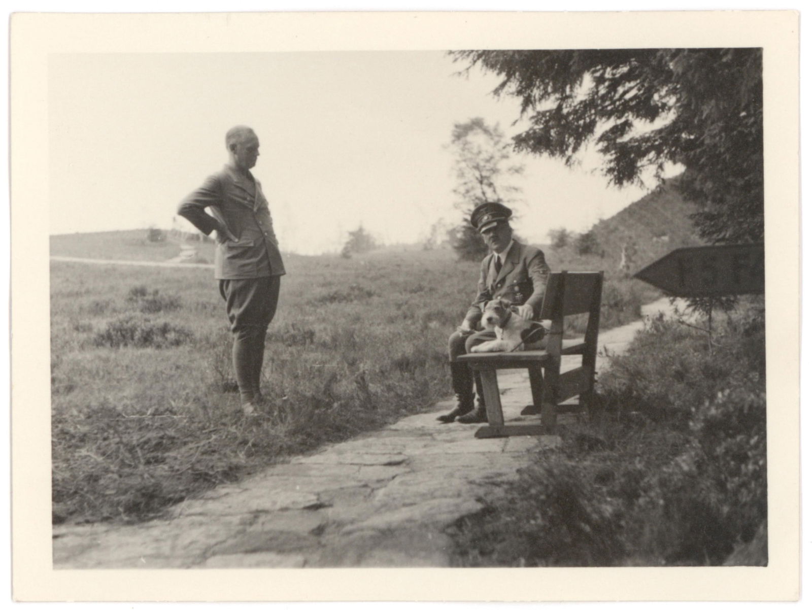 ADOLF HITLER ORIGINAL PHOTOGRAPH BY HOFFMANN: Fine 4.5 x 3.25 in b/w original photograph showing Hitler seated on a bench and petting a dog as an officer stands before him, the image taken by his personal photographer Heinrich Hoffman. Hoffmann's