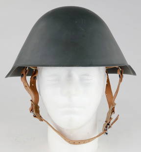 EAST GERMAN ARMY M56 HELMET: An M56 helmet made in Communist East Germany. It bears grey paint on the gently sloping shell. The leather liner has sponge protectors on both the front and back with a leather chinstrap and a plastic