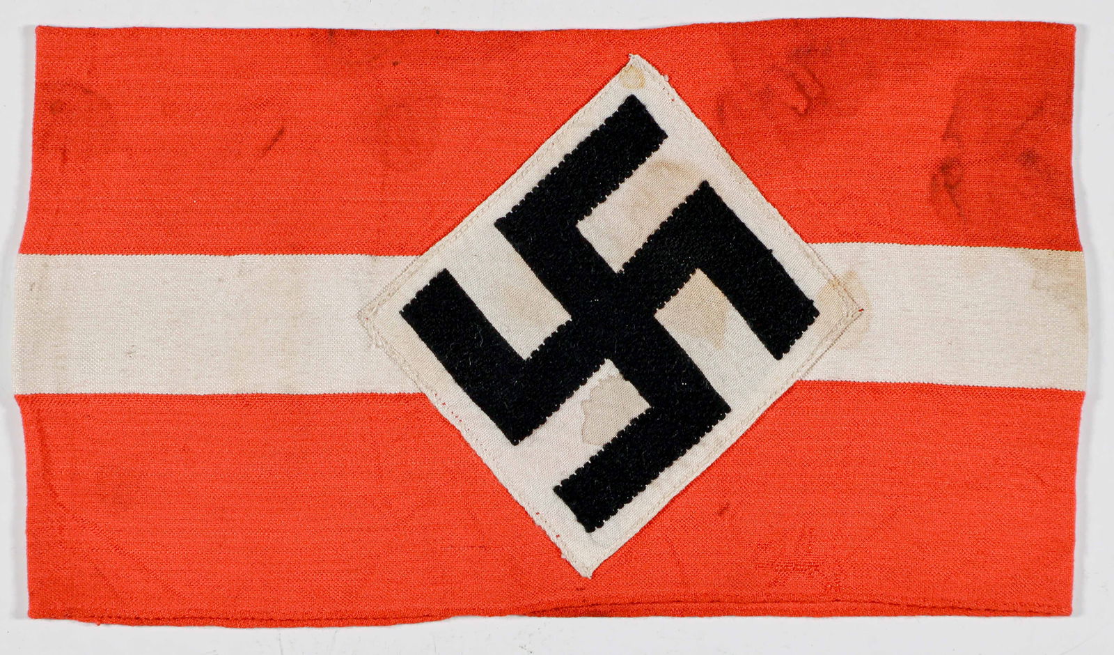 HITLER YOUTH ARMBAND: Red cotton Hitler Youth armband, 15 x 4 in., bearing a white applied band, with a BeVo-style embroidered mobile swastika in the center. Sewn at the ends, shows some brown stains overall, else very goo