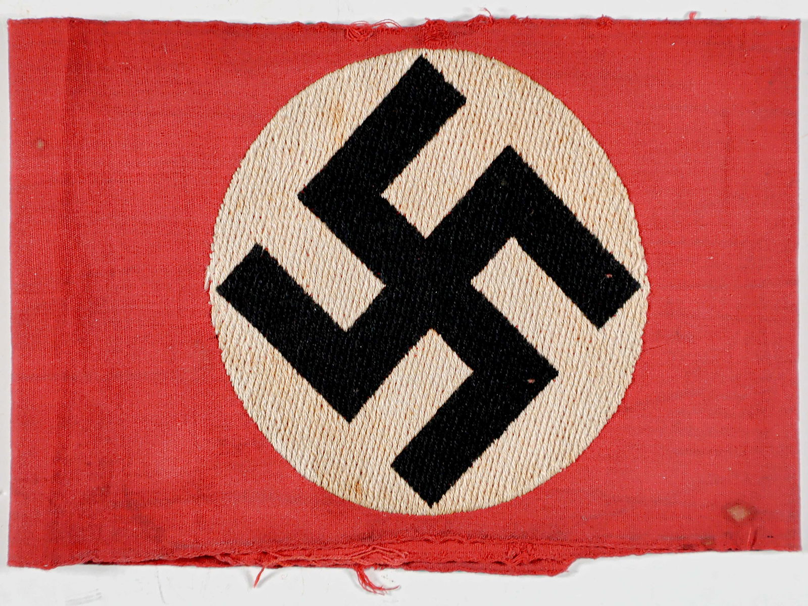 N.S.D.A.P. ARMBAND: Red cotton N.S.D.A.P. armband, 15.5 x 4 in., bearing an expertly embroidered black mobile swastika within a white circle. The ends were sewn, but the stitching has been cut. Frayed at the ends and edg