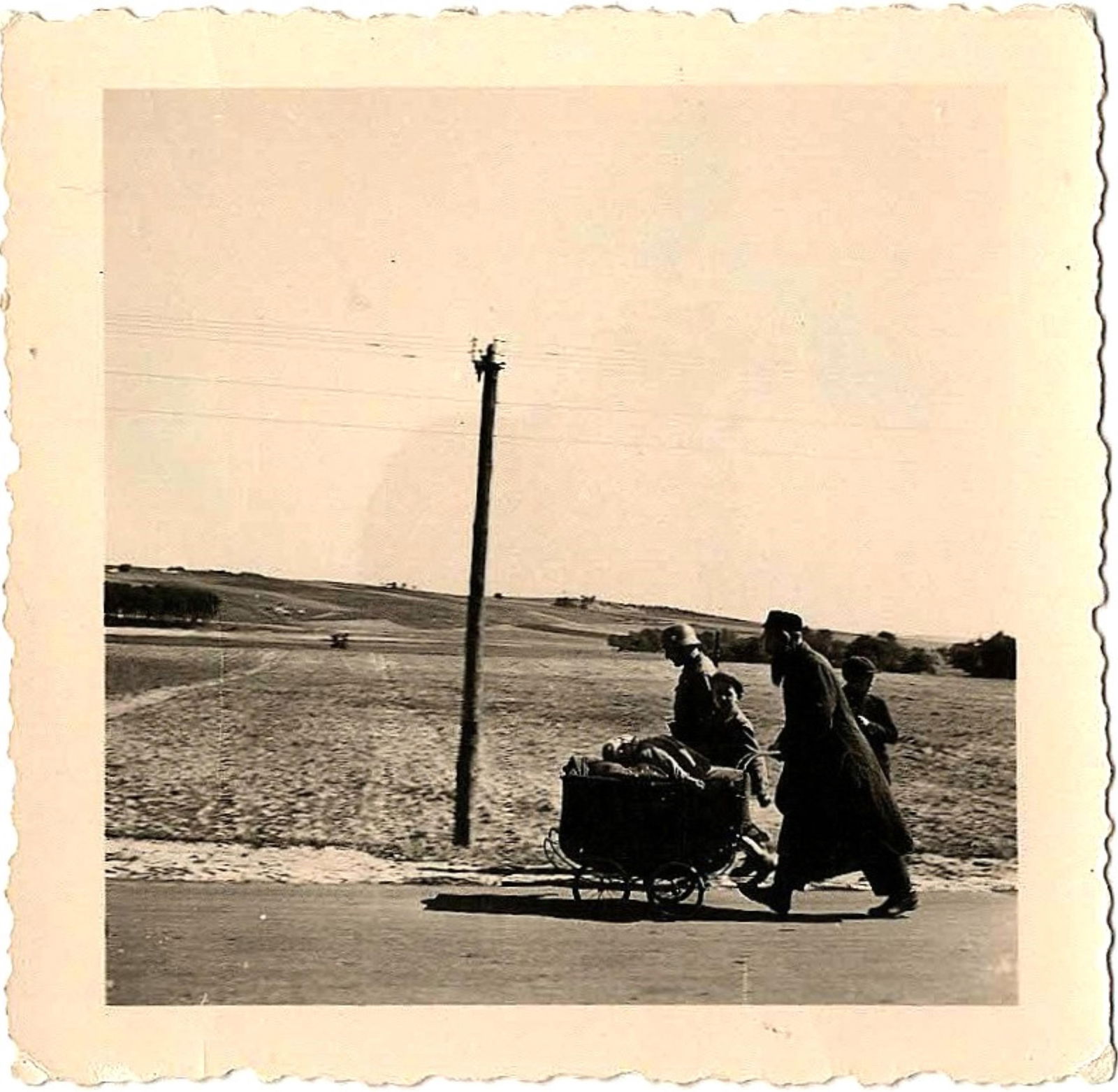 JEW WITH GERMAN GUARD HOLOCAUST PHOTOGRAPH: Unexplainable, gripping original photograph, 2.5 x 2.5 in. b/w, likely taken in Poland ca. 1940. The image shows a tall, bearded Jewish man wearing a long overcoat pushing a baby carriage and taking
