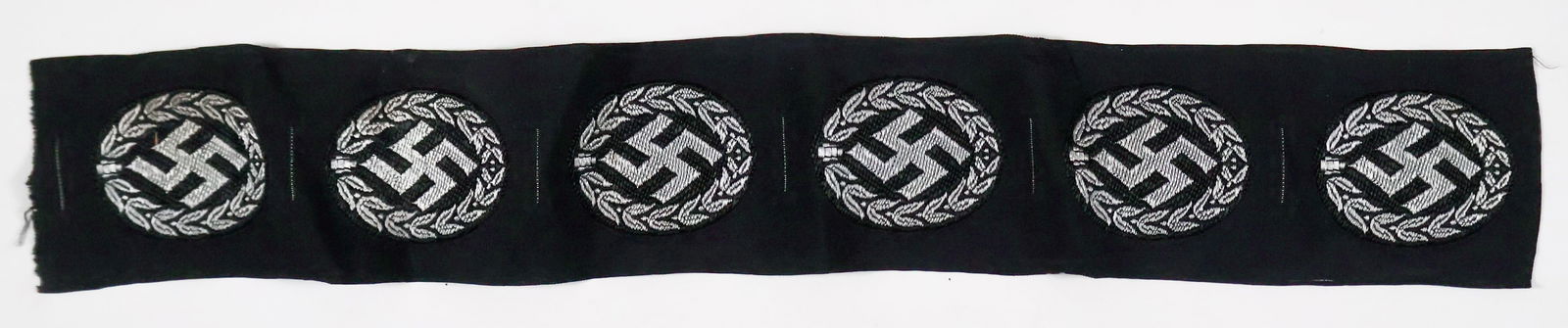 SCHUMA (POLICE) CAP INSIGNIA (6): Lot of six unused, unissued Schuma Sicherheitspolizei (Internal Security Police) cap insignia in a single strip, embroidered silver thread on black backing, fine condition. A good dealer's lot.