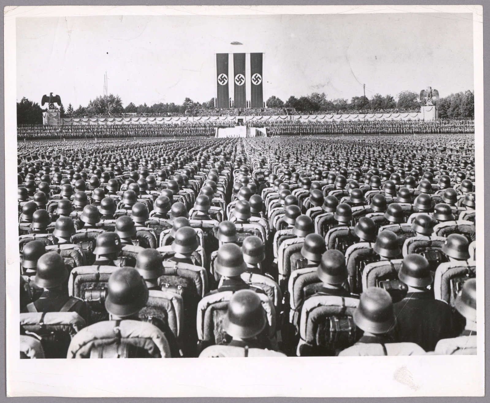 SS TROOPS AT NUREMBERG RALLY: Postwar-issue press photograph, 10 x 8 in. b/w, the famous image of perhaps thousands of SS soldiers listening to Adolf Hitler as he addresses them at the rally grounds at Nuremberg. The verso