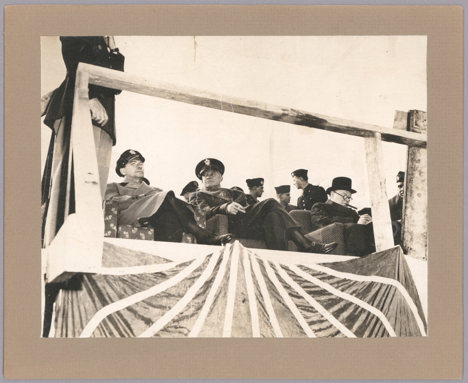 WORLD WAR II PHOTO: EISENHOWER AND CHURCHILL: A 9.25 x 7 in. original sepia photograph showing Dwight D. Eisenhower, cigarette in hand, seated with another American military leader on a podium, while Churchill, cigar clamped in mouth, sits to