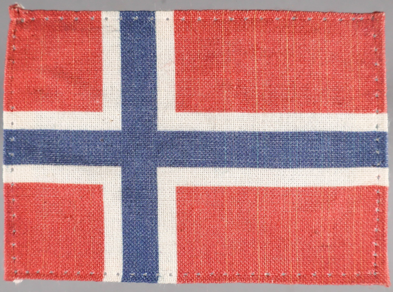 NORWEGIAN WAFFEN-SS VOLUNTEER’S UNIFORM FLAG INSIGNIA: A scarce piece of uniform insignia, a single-sided Norwegian flag patch, 3.25 x 2.25 in., clamp-dyed on cotton bunting and with stitch holes around the edge indicating removal from a uniform. This