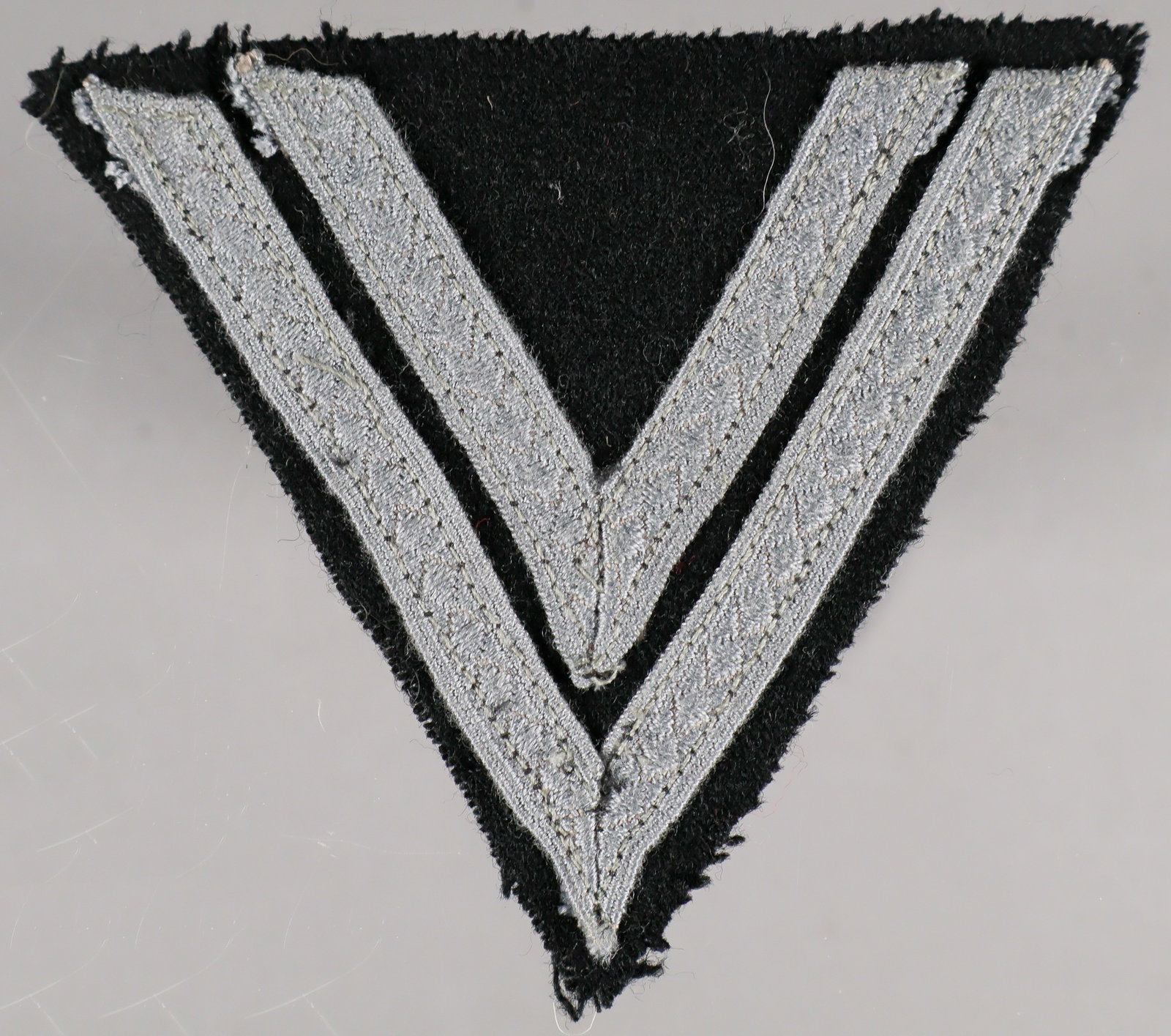 WAFFEN-SS ROTTENFUHRER’S RANK CHEVRON: A Waffen-SS uniform sleeve insignia patch, 3.75 x 3.25 in., consisting of two matte-gray tresse chevrons machine-sewn to a triangular black wool base, indicating the rank of Rottenfuhrer. In very