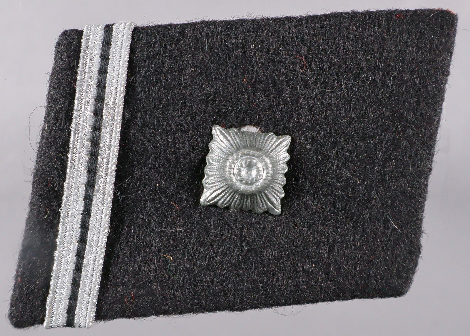 WAFFEN-SS SCHARFUHRER’S COLLAR TAB: A wartime-made Waffen-SS rank collar tab, 2 x 1.5 in., constructed of black wool pasted over a brown buckram core, without piping, fitted with a single metal ‘pip’ over a band of metallic