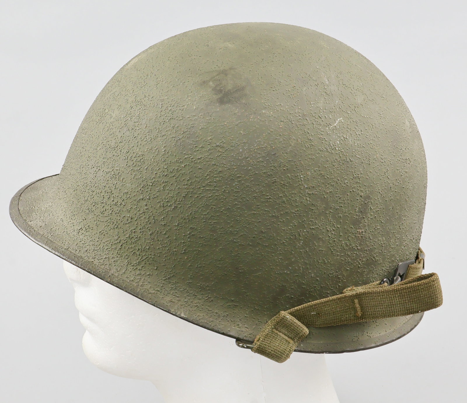 U.S. FRONT SEAM M1 HELMET BY SCHLUETER: Mid-war U.S. steel M1 combat helmet, the exterior finished olive-green with a sand texture, featuring a painted stainless steel rim with seam at the front. The chin strap bales are of the swivel type,