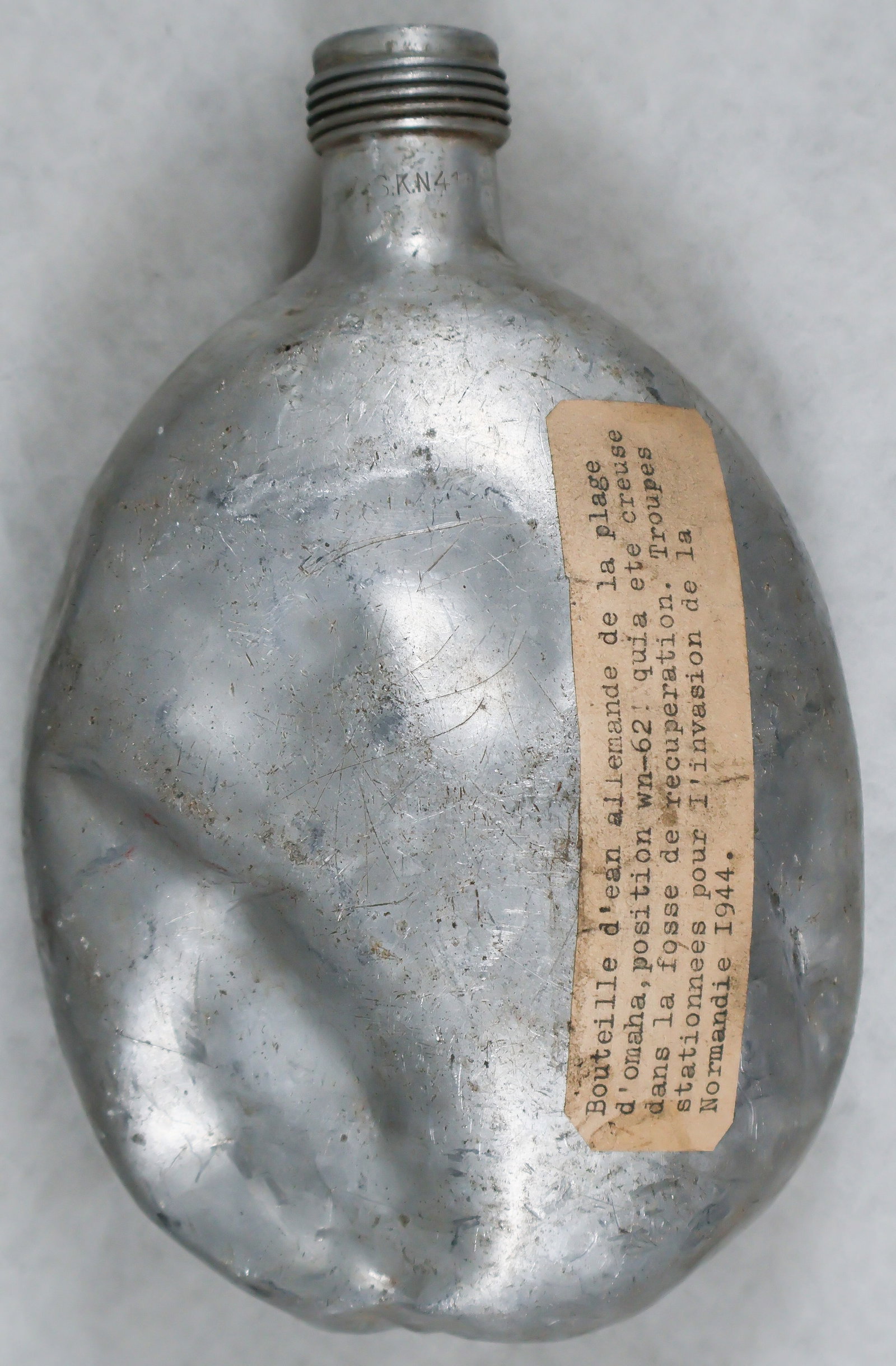 GERMAN CANTEEN RECOVERED AT OMAHA BEACH: German aluminum canteen found along the German defensive line at OMAHA Beach in Normandy. The rather battered canteen bears an old adhesive label with typed provenance in French: 'German water bottle