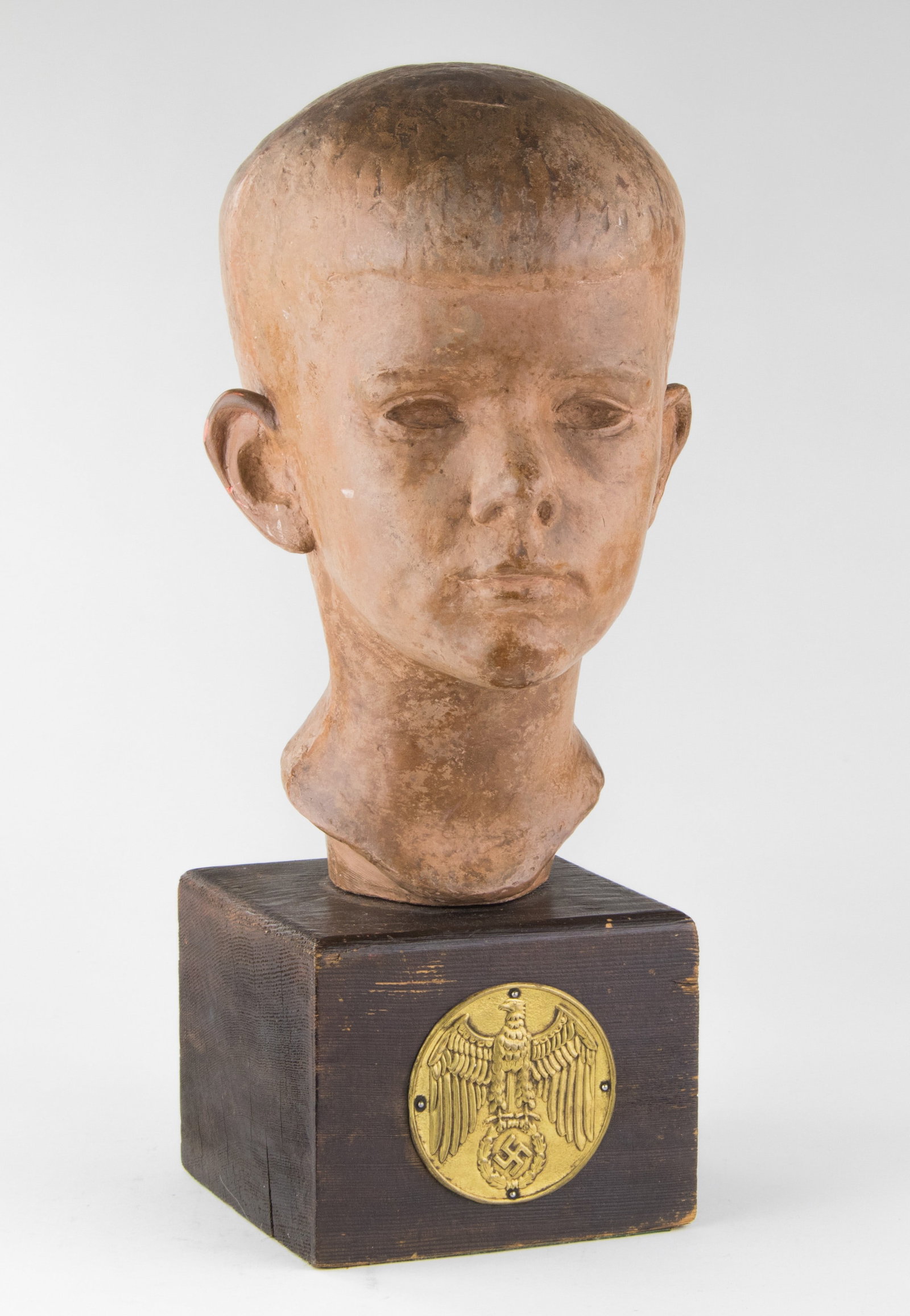 HITLER YOUTH BUST: A ceramic bust depicting a Hitler Youth, 6.5 x 11.75 in., placed on a 4.5 in. wooden base. The front of the base features a gilded plaque featuring the German National eagle, its talons gripping a wre