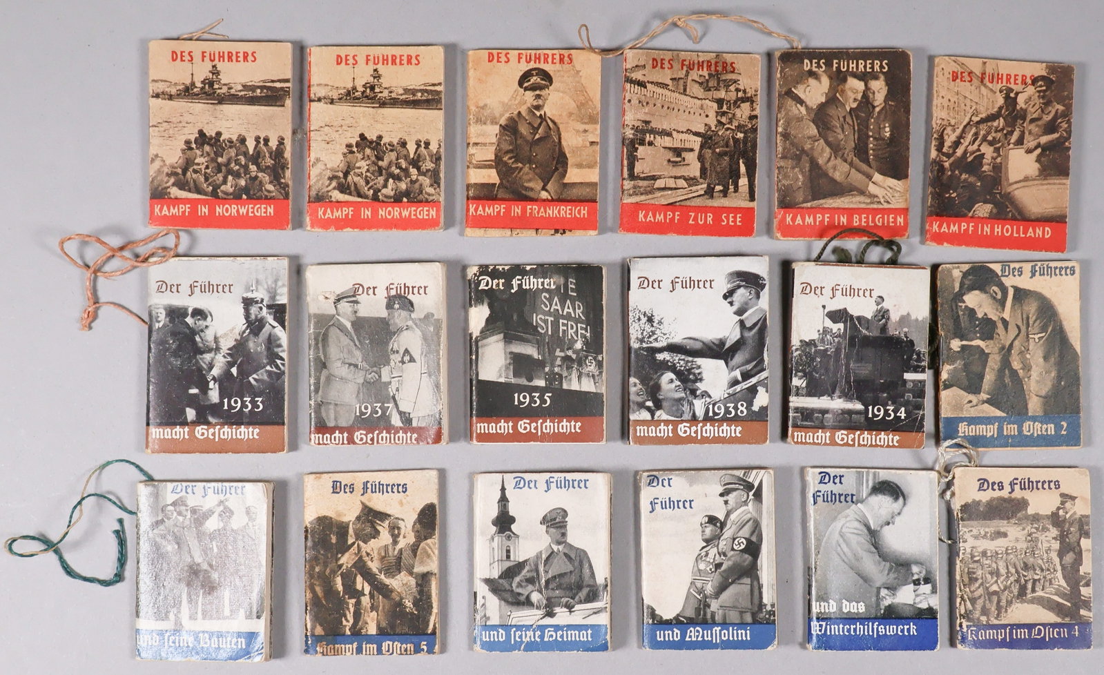 MINIATURE HITLER BOOKS: A collection of 18 miniature Winterhilfswerk books about Hitler, 1.75 in. x 2 in., staple-bound, with some bearing string for hanging at top left. Several bear contemporary tags on verso. Condition