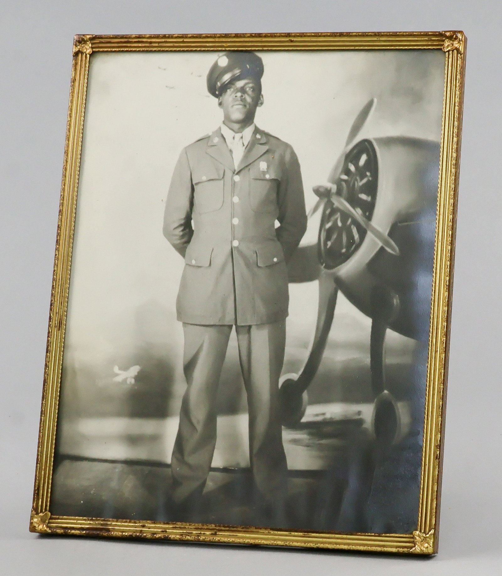 WORLD WAR I BLACK SOLDIER PHOTOGRAPH: Photograph, 8 x 10 in. [sight] b/w, shows a black aviator standing in uniform before a backdrop of a bomber. Set into a gold metal frame, very good. Not examined out of frame.
