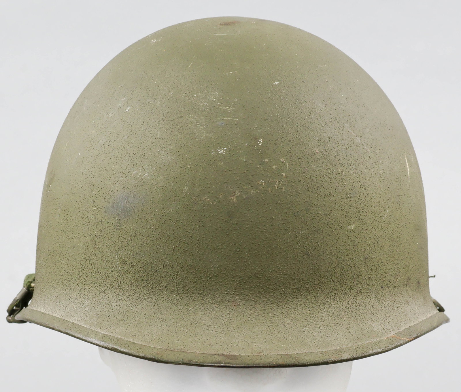 KOREAN WAR-ERA U.S. M1 HELMET: A U.S. steel M1 combat helmet, the exterior finished olive-green with a sand texture, featuring a manganese rim with seam at the rear. The chin strap bales are of the swivel-type, and the olive-green
