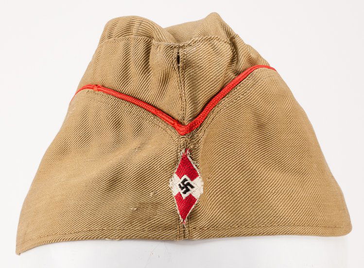 HITLER YOUTH CAP: HITLER YOUTH CAP Hitler Youth cap, 10 «" long in brown cotton twill with red piping, an embroidered H.K. emblem sewn to the front. A remnant of white tag also remains within. Very good.