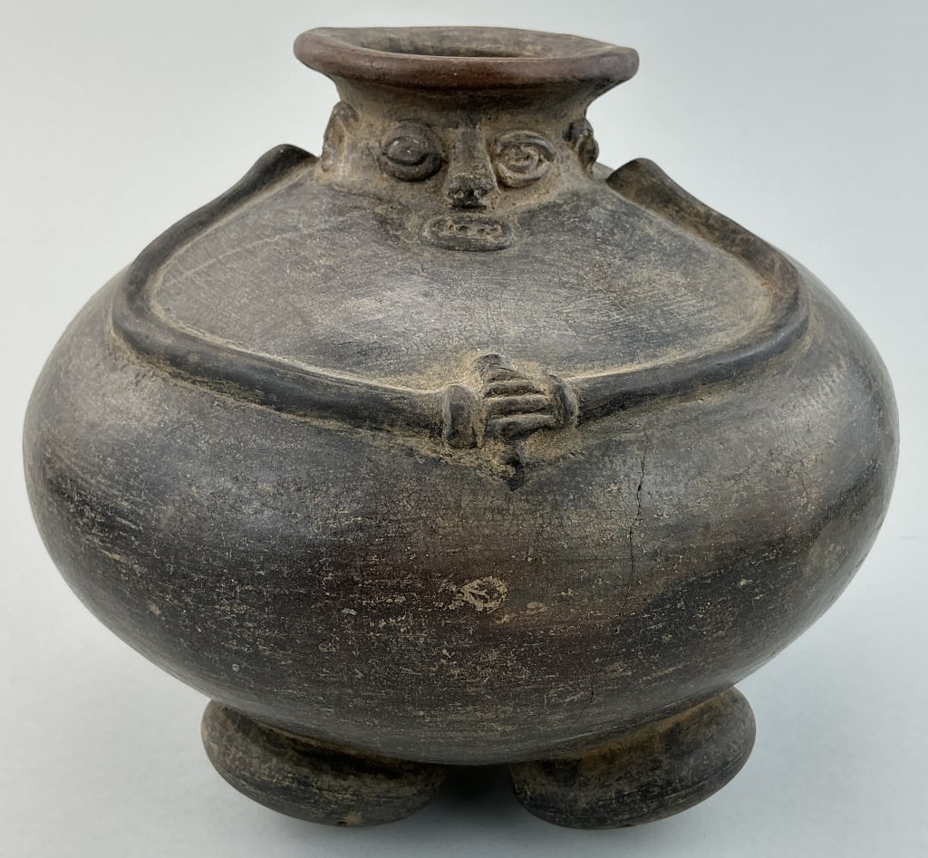 Pre-Colombian Chimu Blackware Pottery Vessel.: Measures 9 inches high. North Coast, Peru, Chimu culture. Stylized human head flared spout, bulbous body, hands clasped on the torso and triple bun feet. Condition: Fair with crazing and a vertical cr
